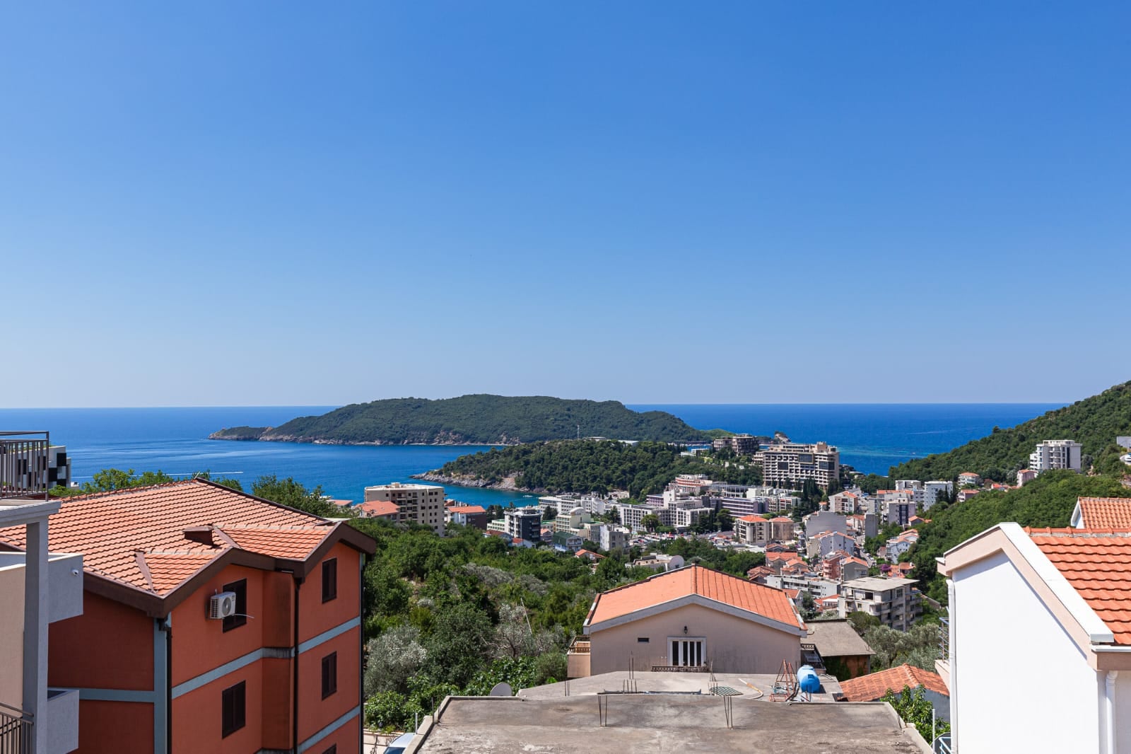 Panoramic view of the sea and the resort of Becici, Montenegro, from a villa terrace with tiled roofs surrounded by greenery.
