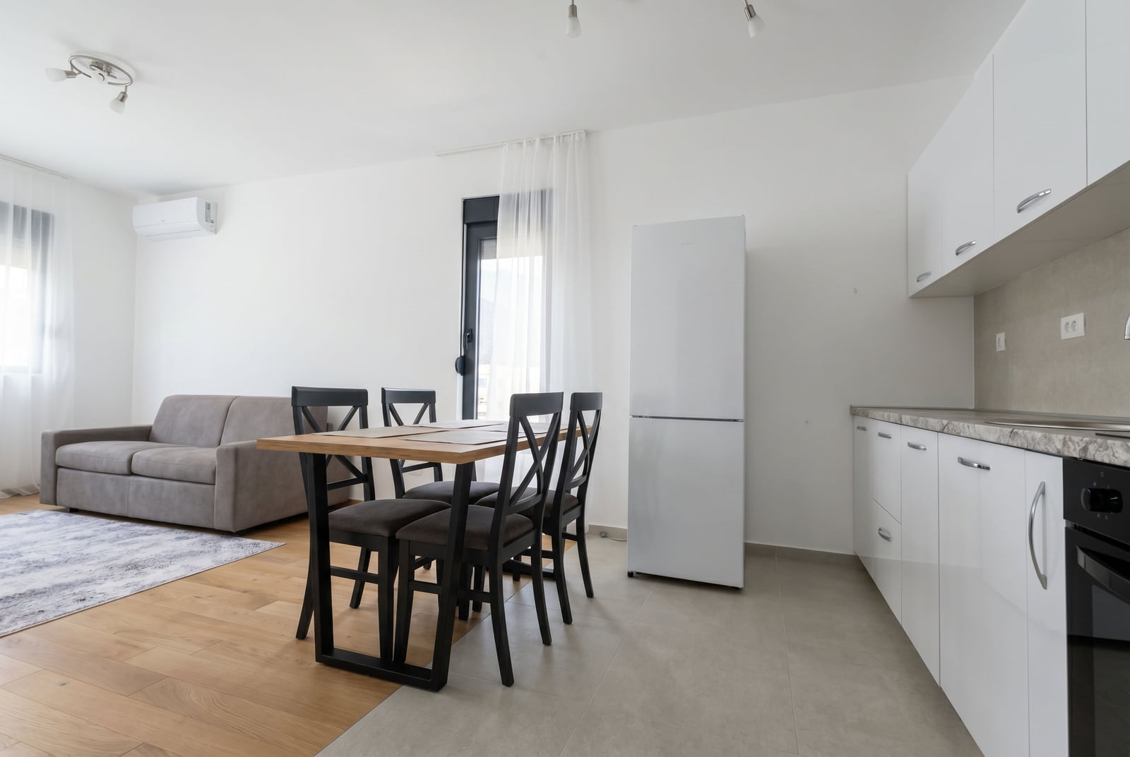 Modern living room with an open-plan kitchen in a Becici apartment, Montenegro: white furniture, dining area, and a grey sofa.