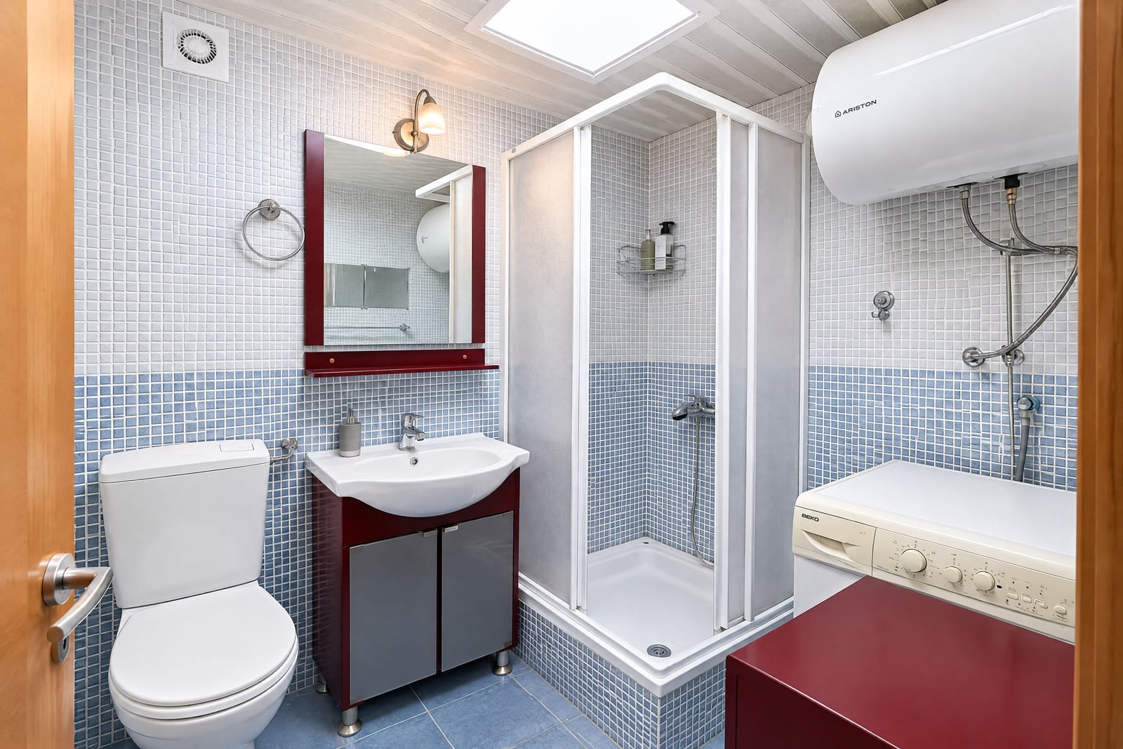 Bathroom with toilet, sink, shower enclosure and washing machine on blue mosaic tiles under a skylight.