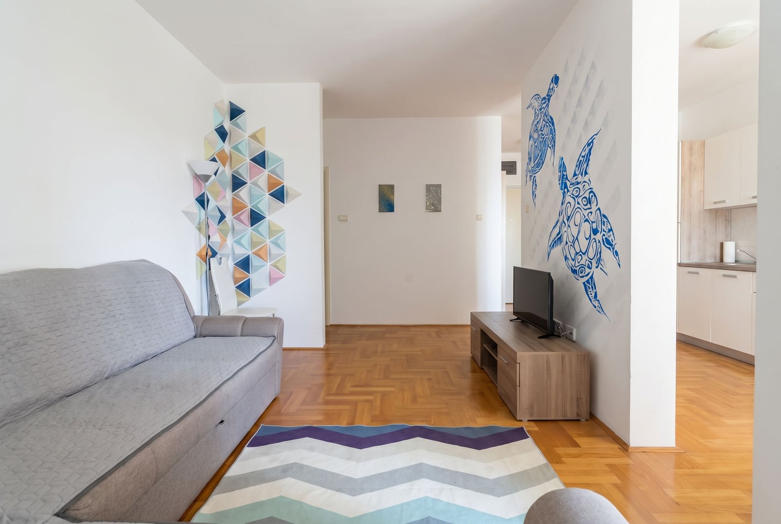 Living room with a gray sofa, parquet flooring, and wall decor in an apartment in Bečići, Budva, Montenegro. View of the kitchen.