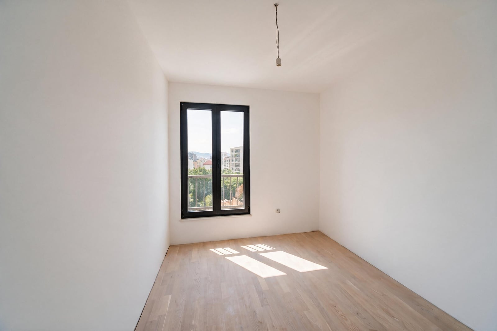 Empty room with white walls, light wood flooring, and a black-framed window overlooking a balcony and city.
