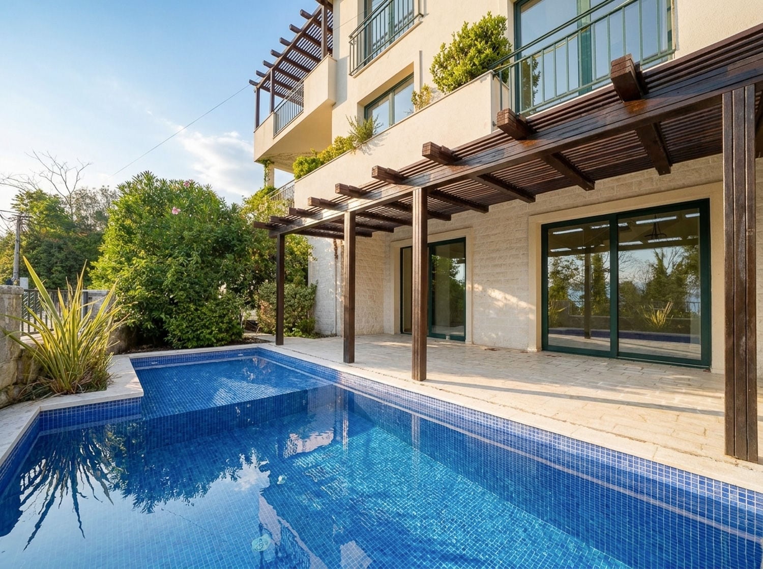 Blue tiled swimming pool adjacent to a stone patio, wooden pergola, and lush greenery outside a modern building.
