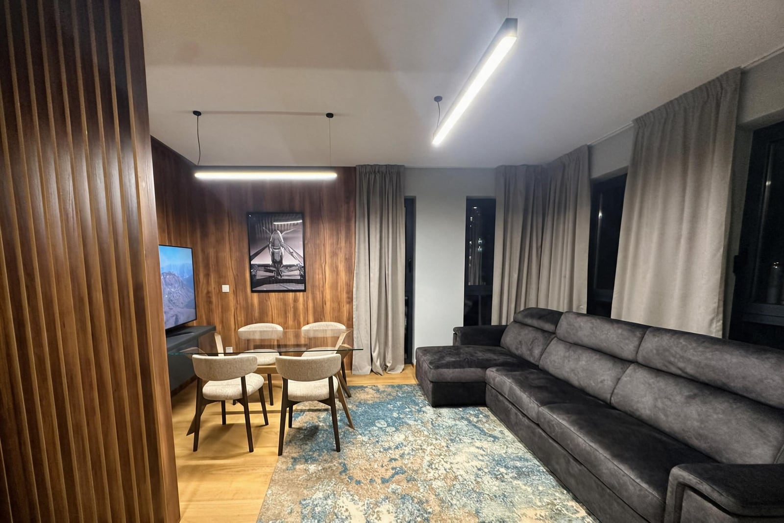 Living room with grey sofa, glass dining table, wood accent wall, and TV.