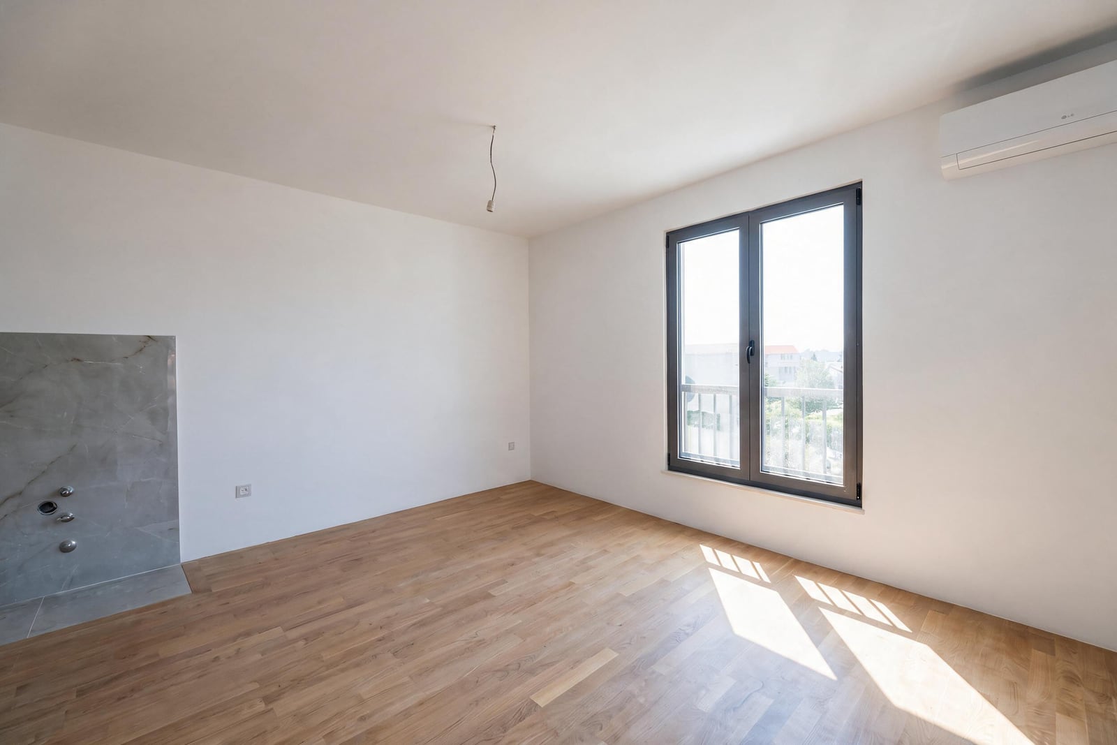 Empty room with wood flooring and a window leading to a balcony. A grey tiled wall with plumbing fixtures is visible on the left.