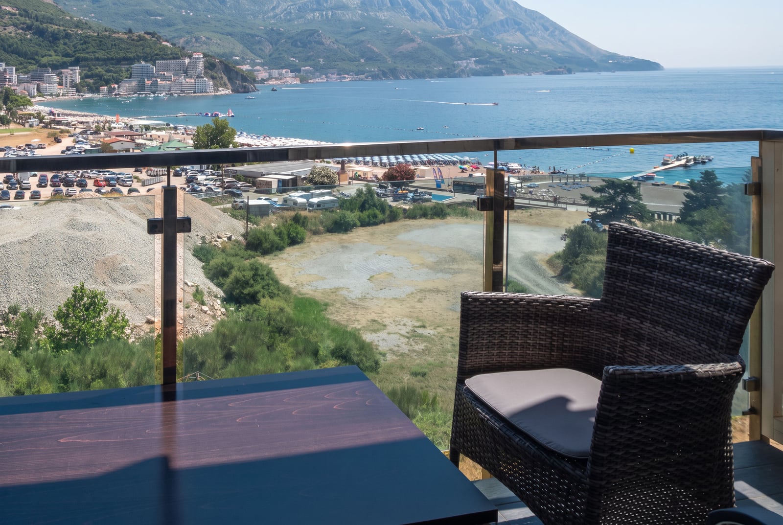 Balcony with wicker chair and table overlooking the sea, beach, and mountains through a glass railing.