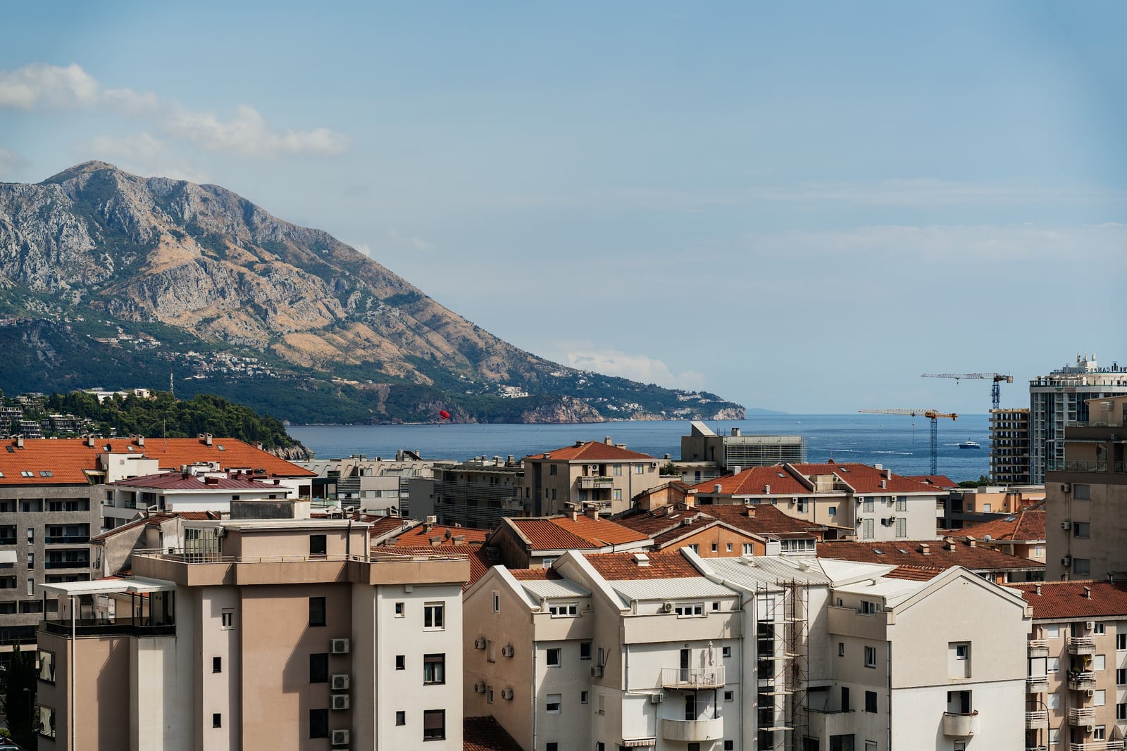 Panoramic view of city rooftops, sea, and a large mountain under a clear sky.