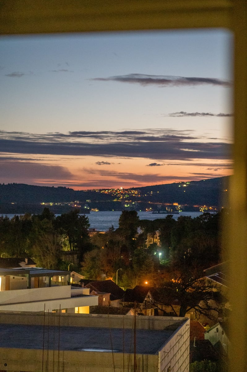 Panoramic view of Tivat and the Bay of Kotor at night, seen from a Montenegrin apartment window, featuring city lights and surrounding mountains.