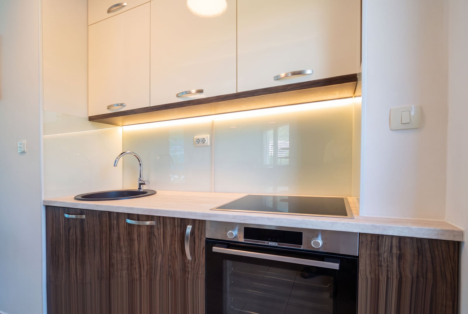 Modern kitchen featuring white upper cabinets, wood-grain lower units, a black sink, and a built-in Bosch oven under warm LED lighting.