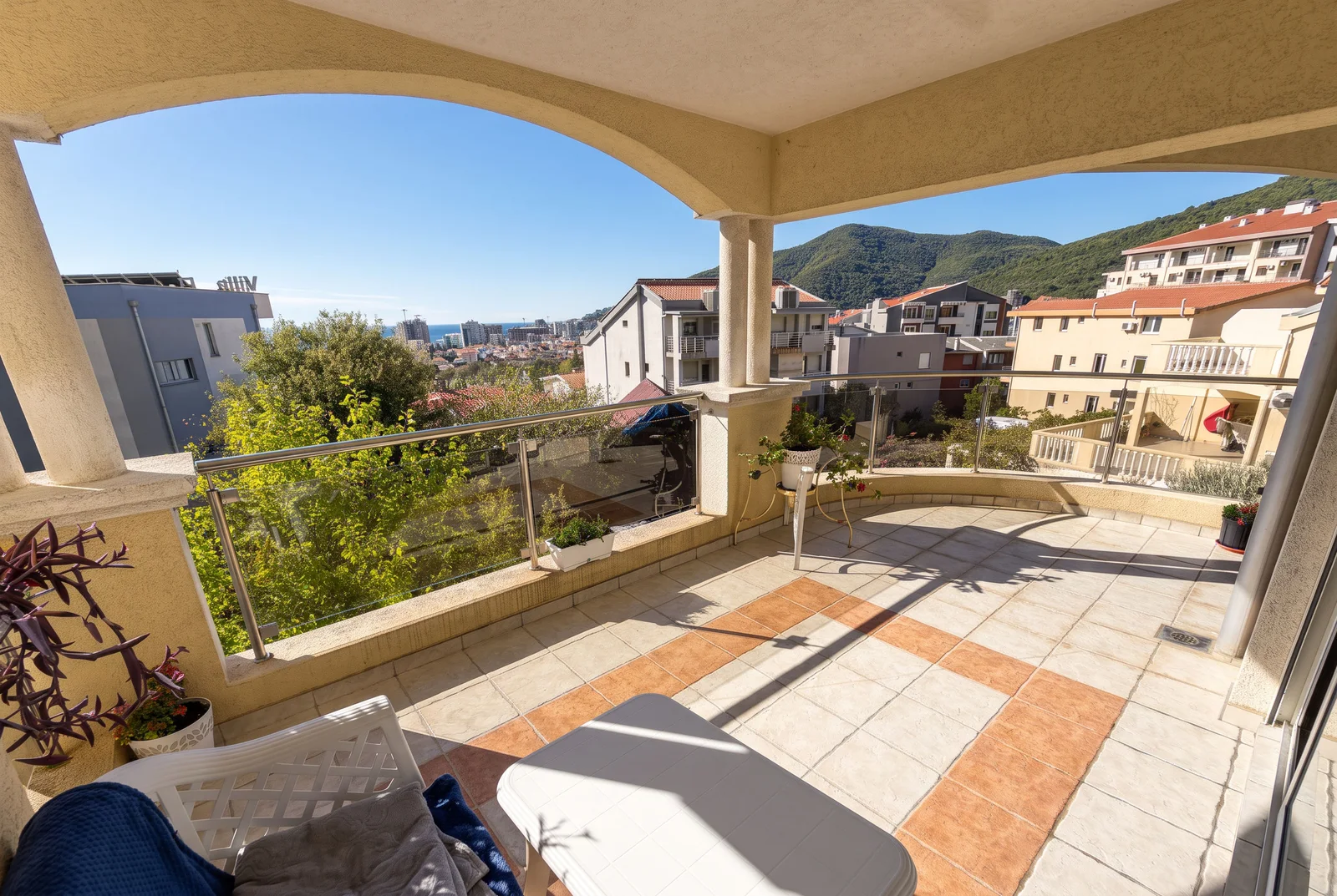 Wide tiled balcony with glass railing overlooking a city and mountains under a blue sky.