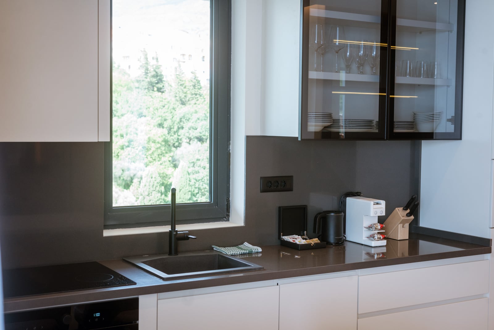 Modern kitchen in an apartment in Bečići, Montenegro: dark countertop, built-in appliances, and a view of greenery from the window.
