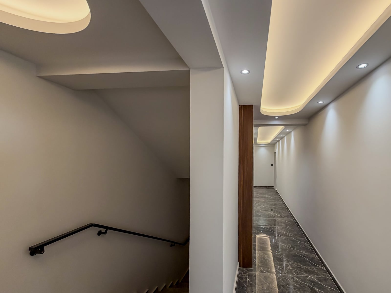 Modern hallway with marble flooring, wood column, and recessed ceiling lighting. A staircase with a black handrail is visible on the left.