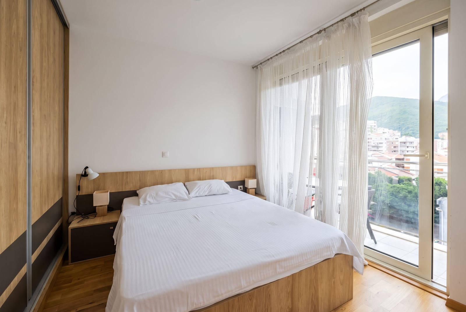 Modern bedroom featuring a wooden bed with white linens, a wardrobe, and a large window with sheer curtains.