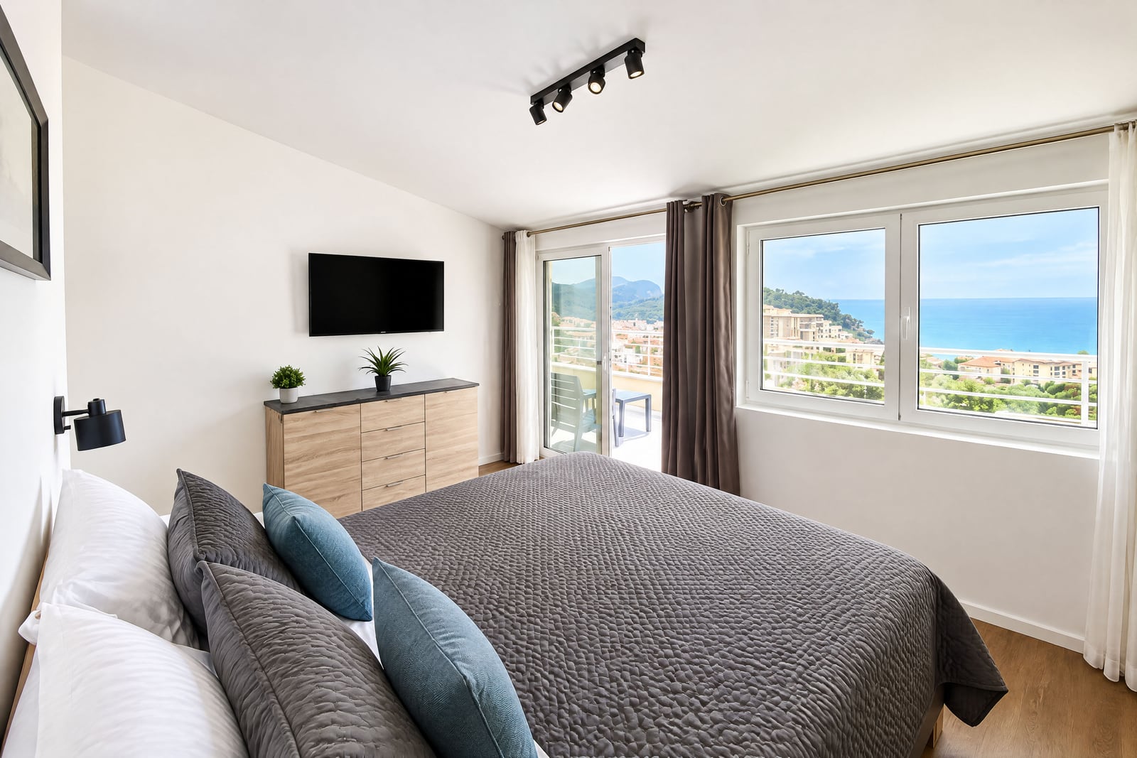 Modern bedroom with a large bed, wall-mounted TV, dresser, and ocean view through windows and balcony door.