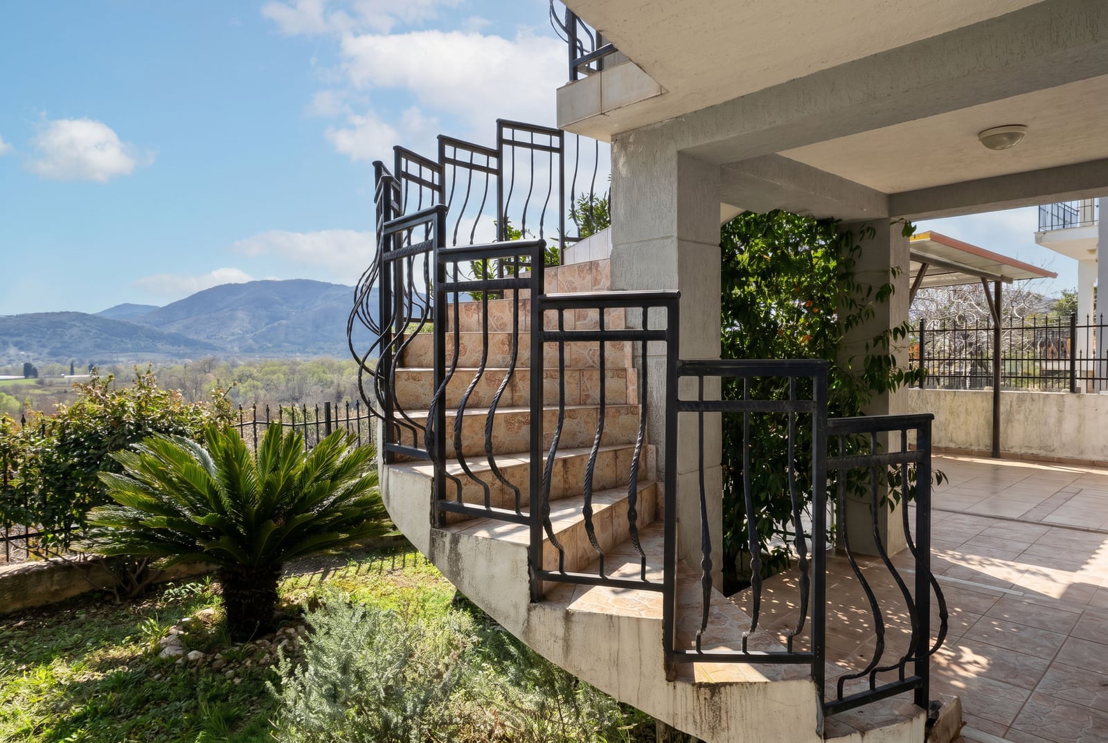 Exterior spiral staircase with wrought iron railings at a house in Buljarica, Montenegro, featuring views of the mountains and a green garden.