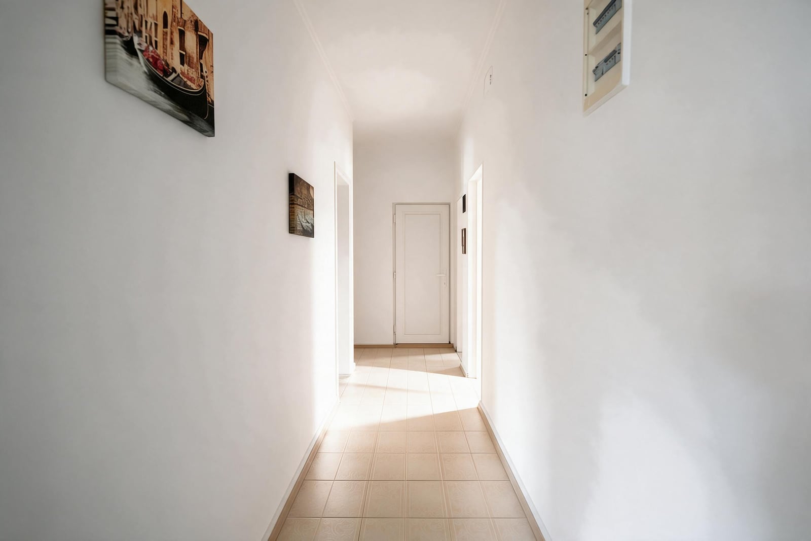 Long white hallway with tiled flooring, wall art, and an electrical panel on the right wall.