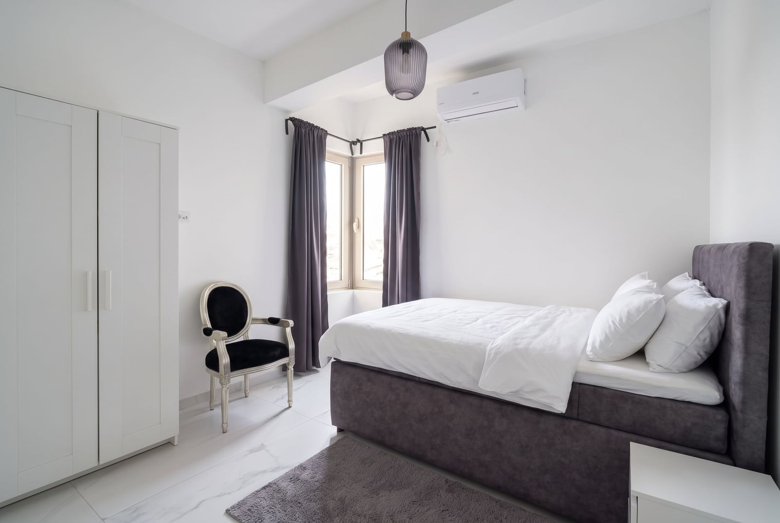 Bright bedroom featuring a grey bed, white wardrobe, black chair, window with curtains, and AC unit.