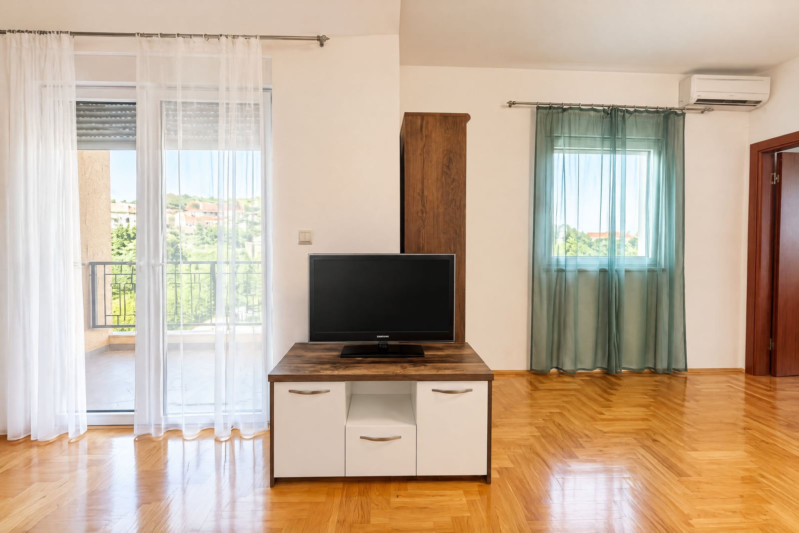 Living room with parquet floor, TV on a stand, balcony door, and wall AC unit.