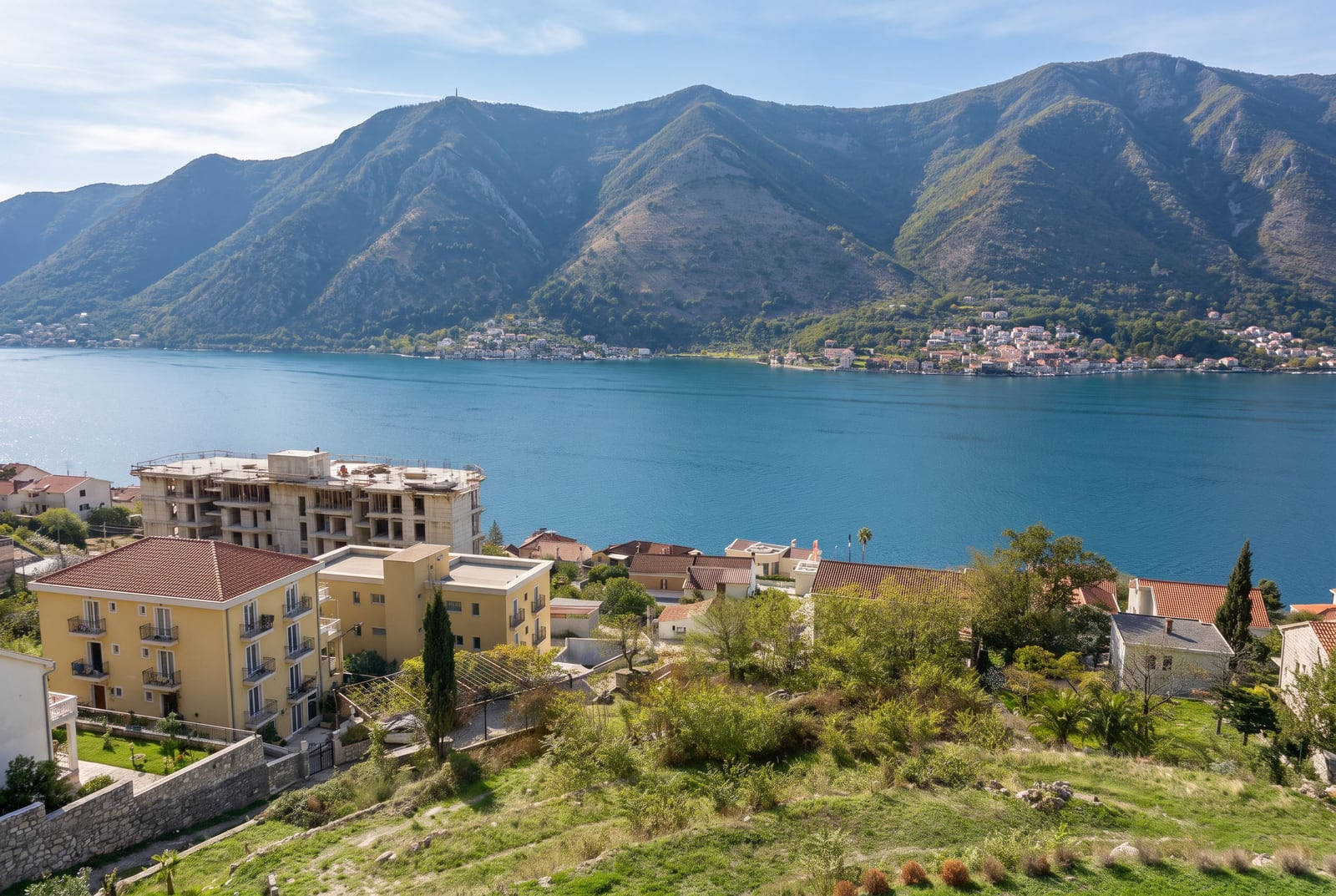 Panoramic view of a blue bay, mountains, and coastal houses with green vegetation in the foreground.