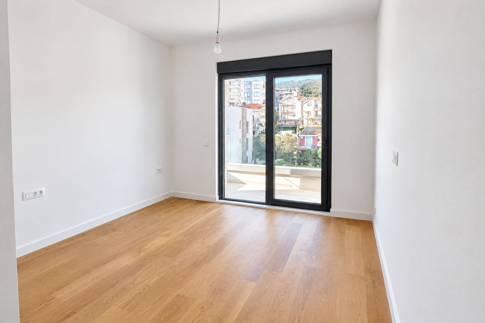 Empty room with wood flooring and white walls, sliding door leads to a balcony overlooking buildings.