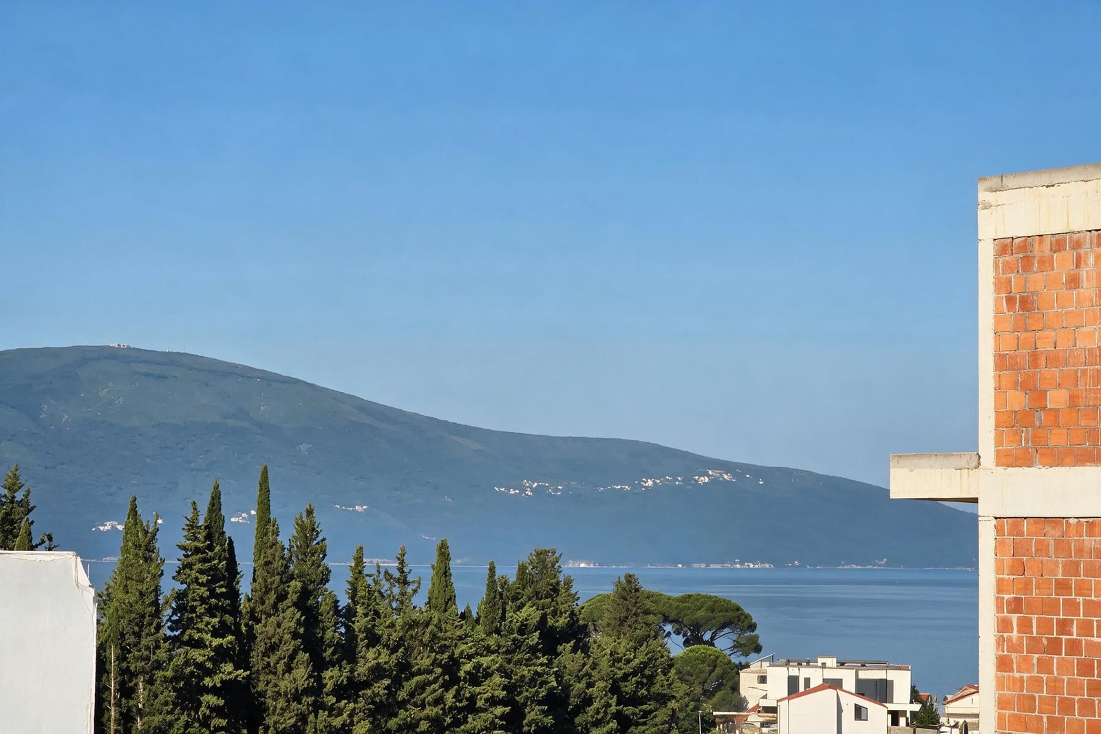View of the sea and mountains with a brick wall, trees, and clear blue sky.