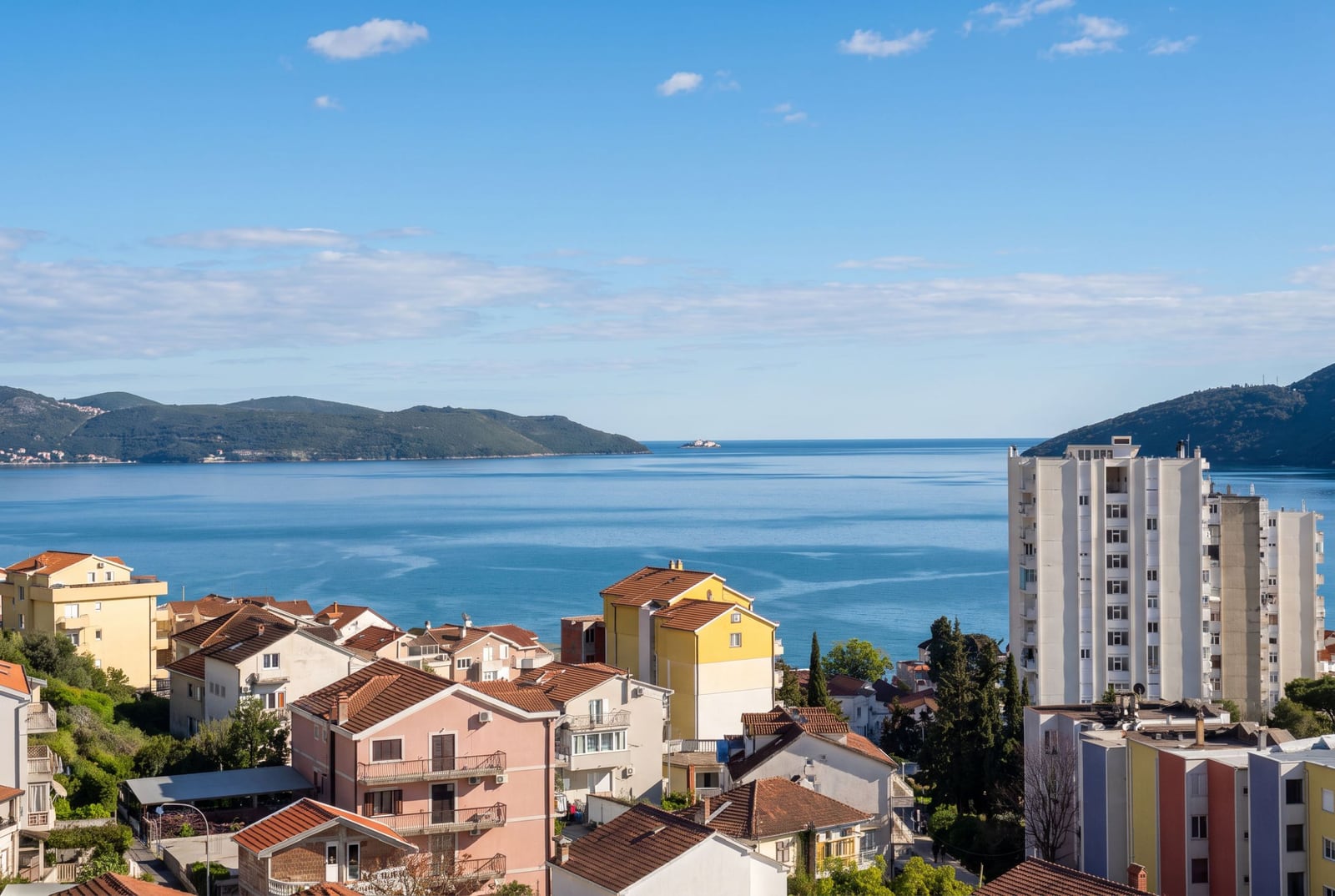 Panoramic view of a blue sea bay, coastal houses with tiled roofs, and green hills under a clear sky.