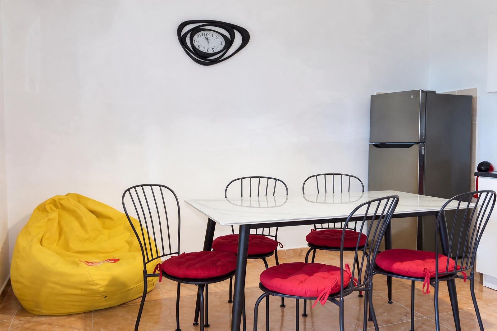 Dining area with white table, black chairs with red cushions, yellow beanbag, and silver LG refrigerator against a white wall.