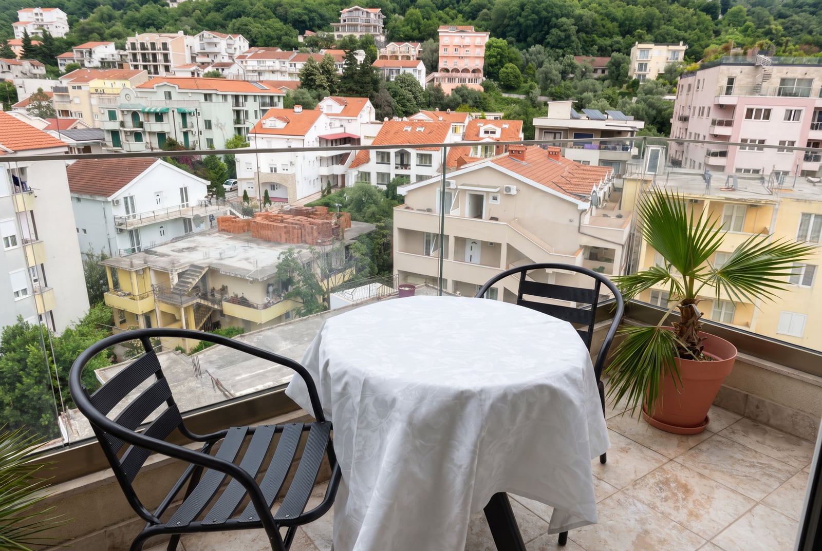 Balcony with black table and chairs under a white cloth, overlooking residential houses and green hills.