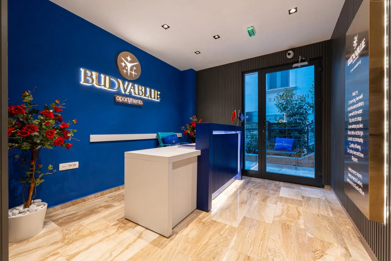 Apartment lobby reception with blue wall, BUDVABLUE logo, white desk, and artificial roses.