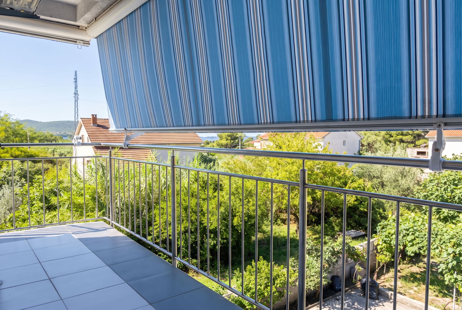 Tiled balcony with stainless steel railing and blue striped fabric awning overlooking greenery.