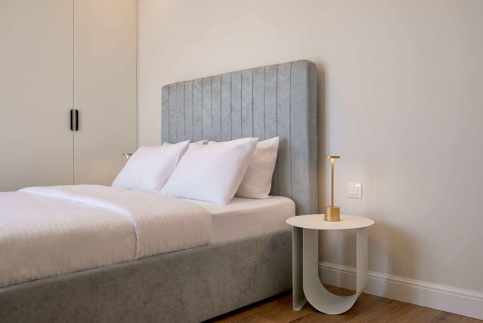 Modern bedroom featuring a grey upholstered bed with white linens, a minimalist side table, and a gold lamp against a plain wall.