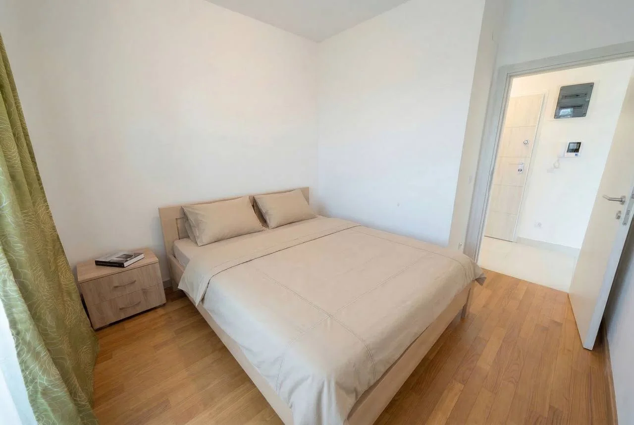 Bedroom featuring a double bed with beige bedding, a wooden nightstand, green curtains, and light wood flooring.