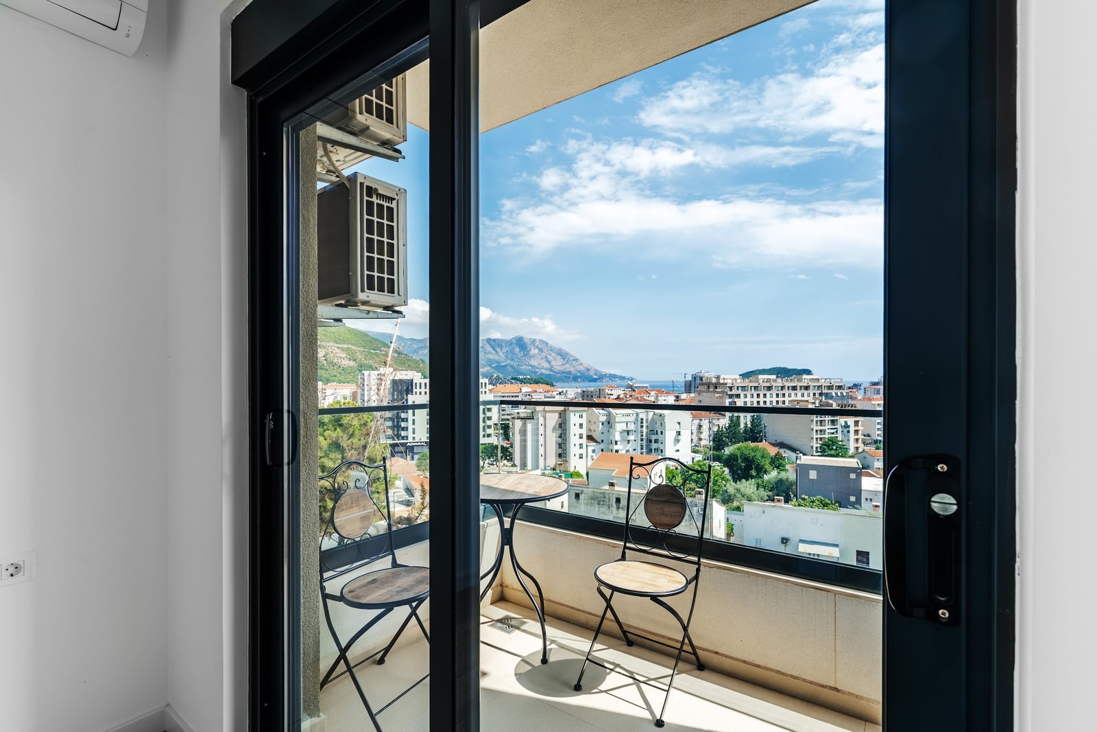 View from an interior room through glass doors onto a balcony with a small table and two chairs overlooking a city and mountains.