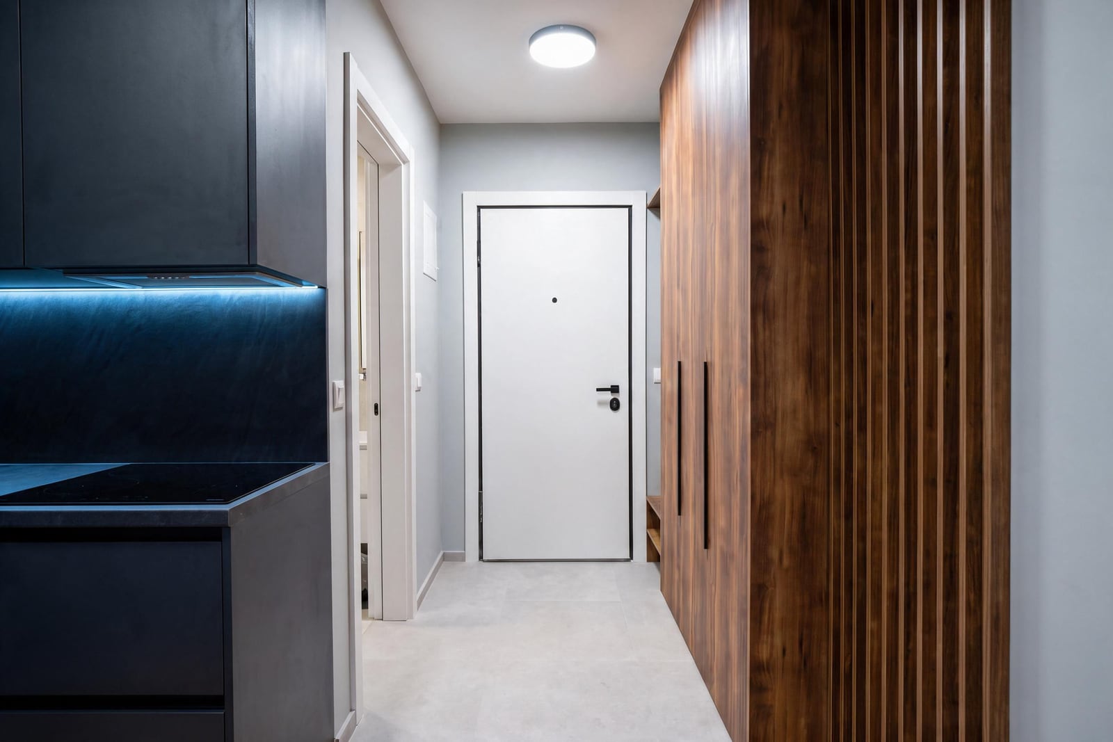 Hallway with white entrance door, wooden wardrobe, and partial view of a dark kitchen with blue under-cabinet lighting.