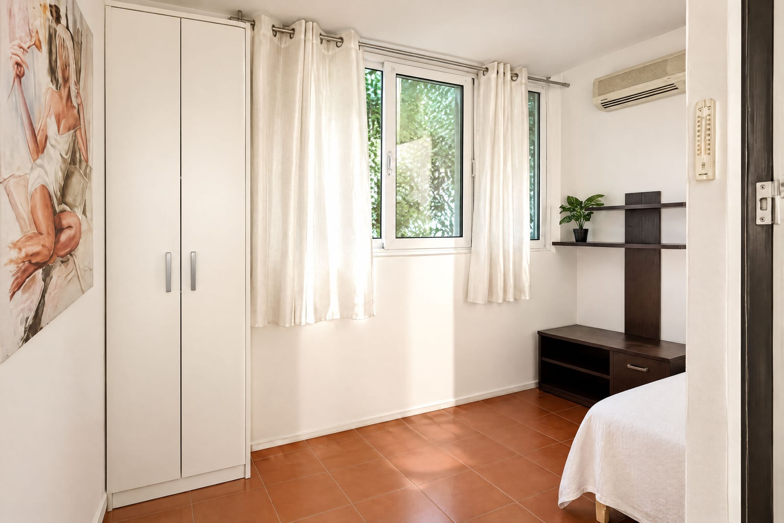Bedroom with white wardrobe, wooden shelf, window with curtains, and edge of a bed on terracotta floor.