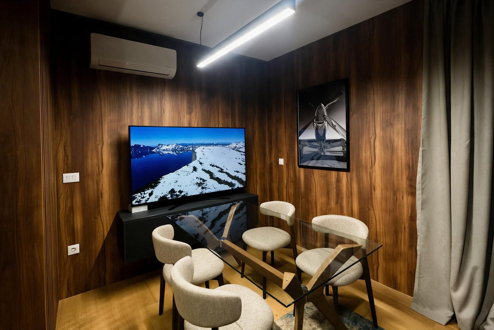 Modern room with wood paneled walls, a glass dining table, four beige chairs, and a large TV on a black console under linear lighting.