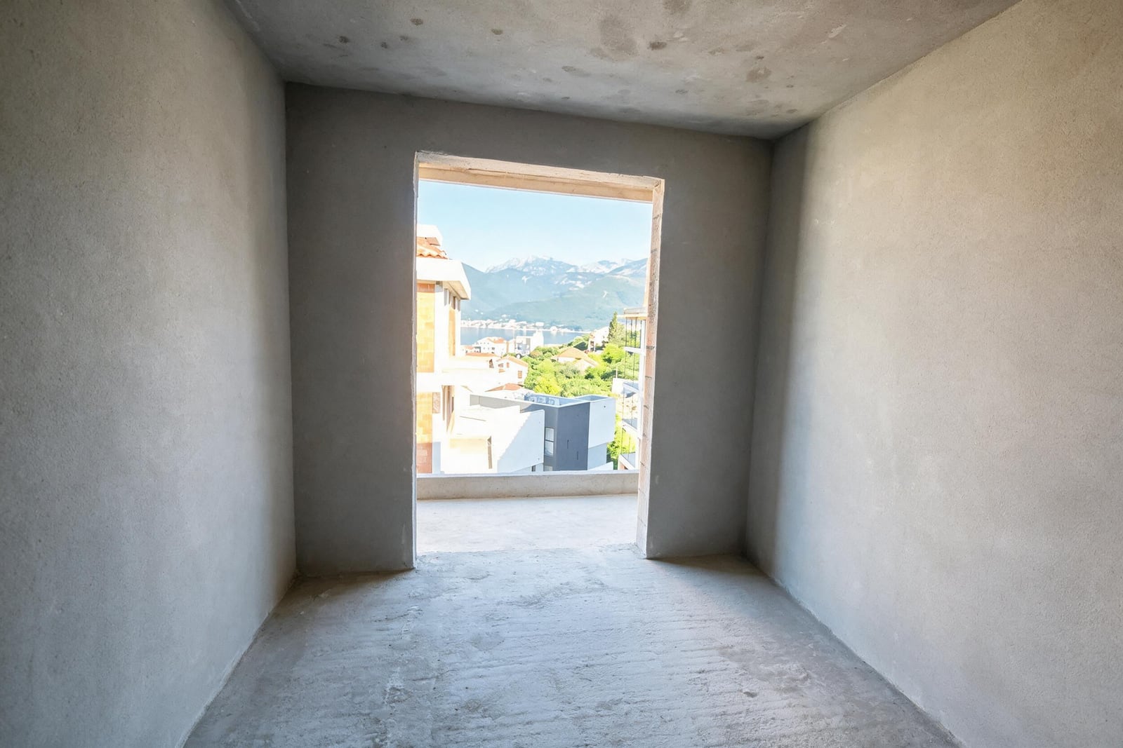 Unfinished room with bare concrete walls and a doorway opening to a terrace overlooking mountains and water.
