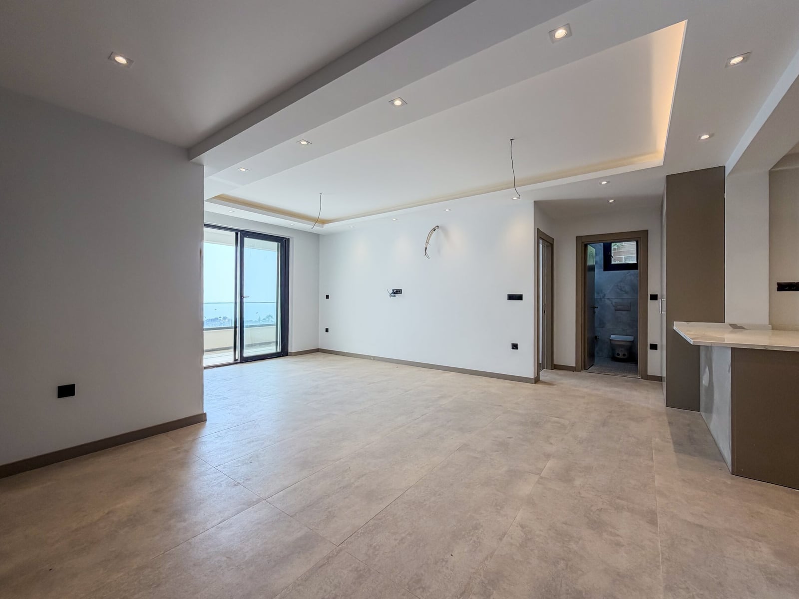Empty room with grey tiles, multi-level ceiling, and balcony door. A kitchen counter edge is visible on the right side.
