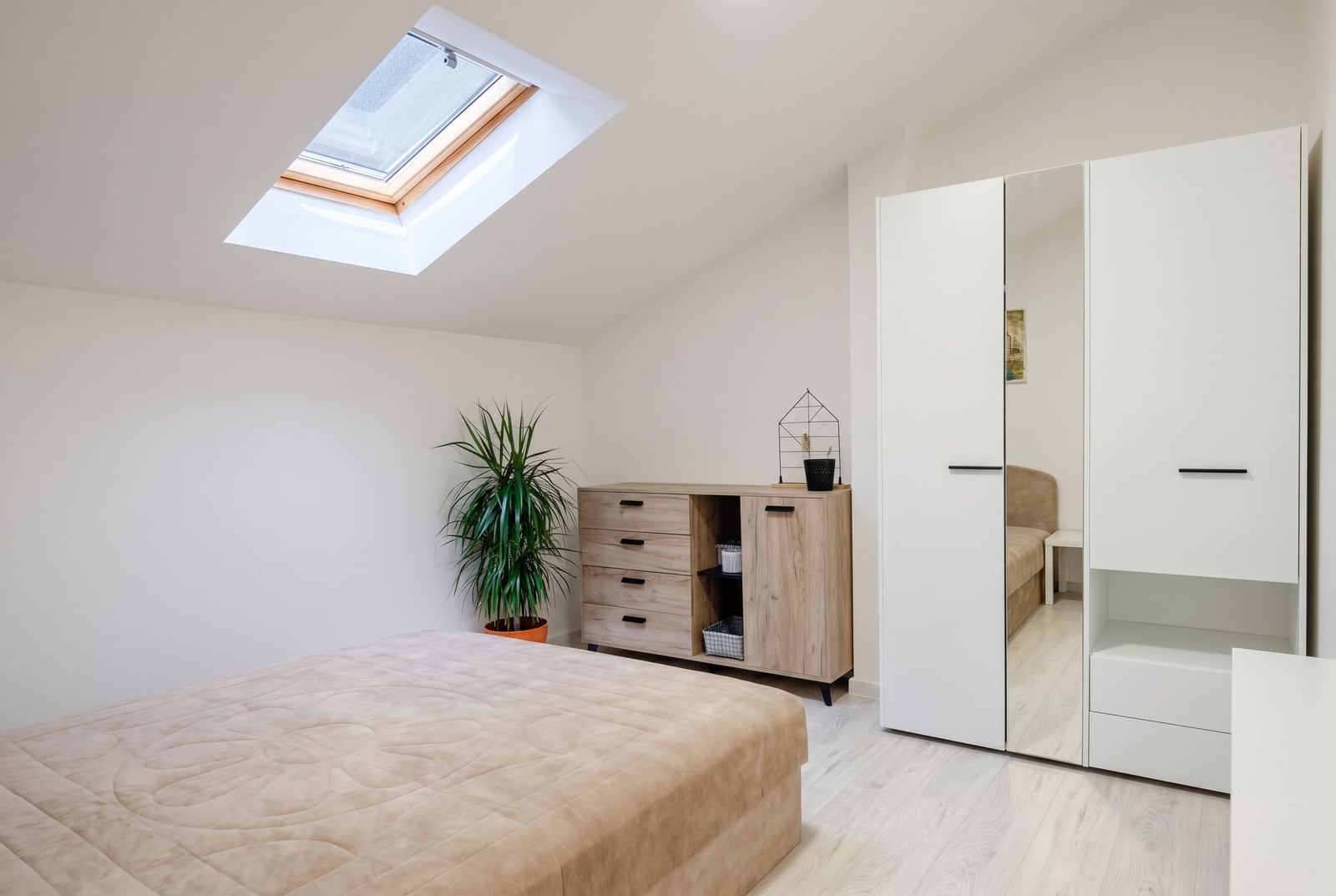 Bright attic bedroom in a Budva apartment, Montenegro: white furniture, mirrored wardrobe, dresser, and skylight.