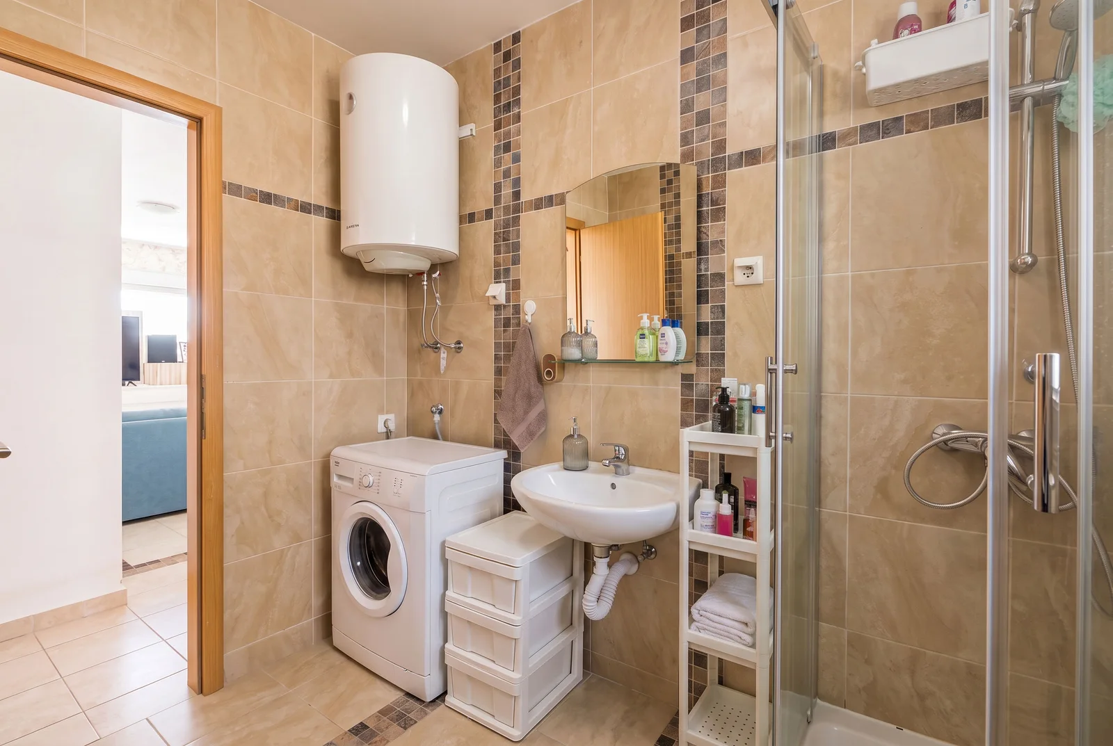Bathroom with shower enclosure, sink, washing machine, and wall-mounted water heater on beige tiles.