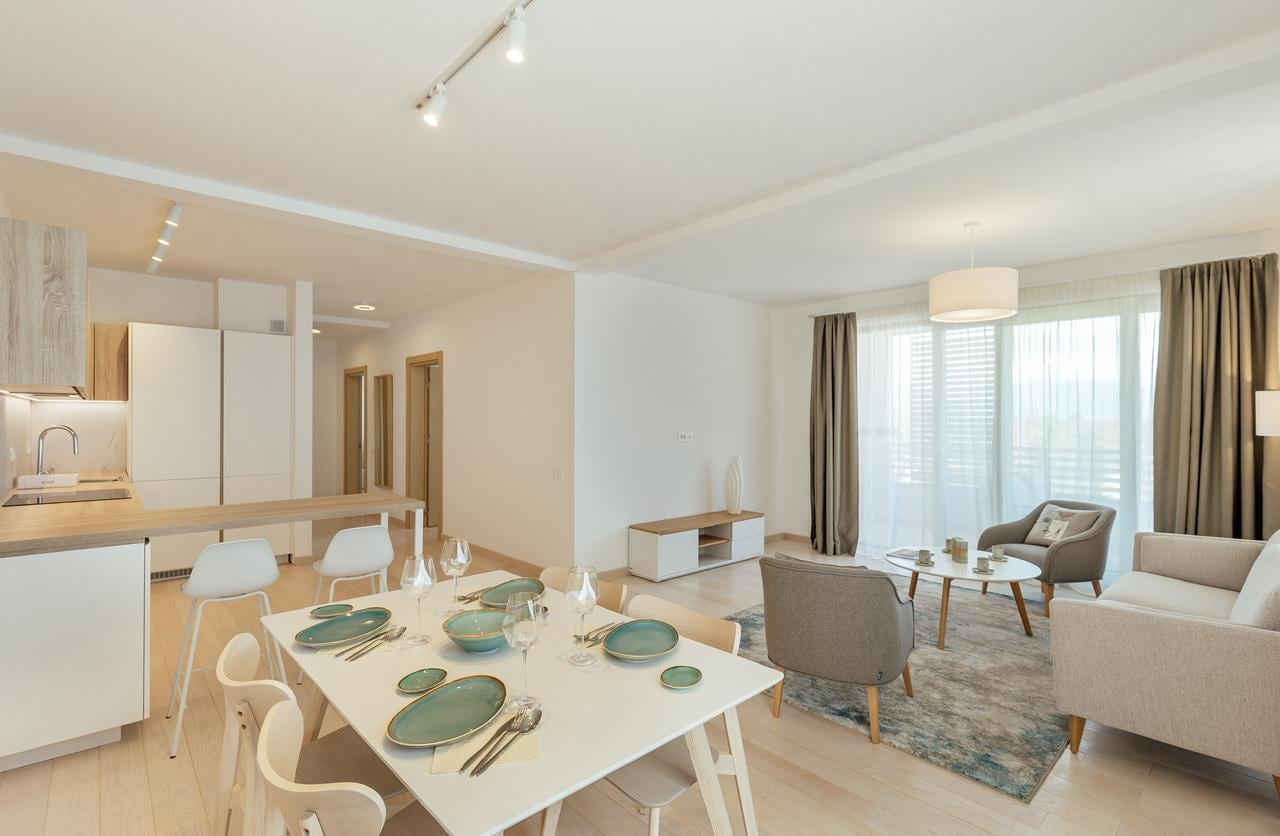 Modern living room with kitchen in an apartment in Igalo, Montenegro: dining area, light-colored furniture, and panoramic windows.