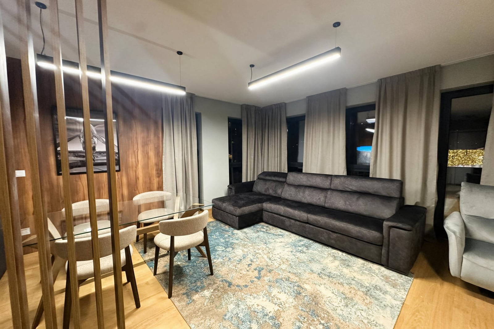 Modern living room featuring a gray sectional sofa, glass dining table, and wooden slat partition.