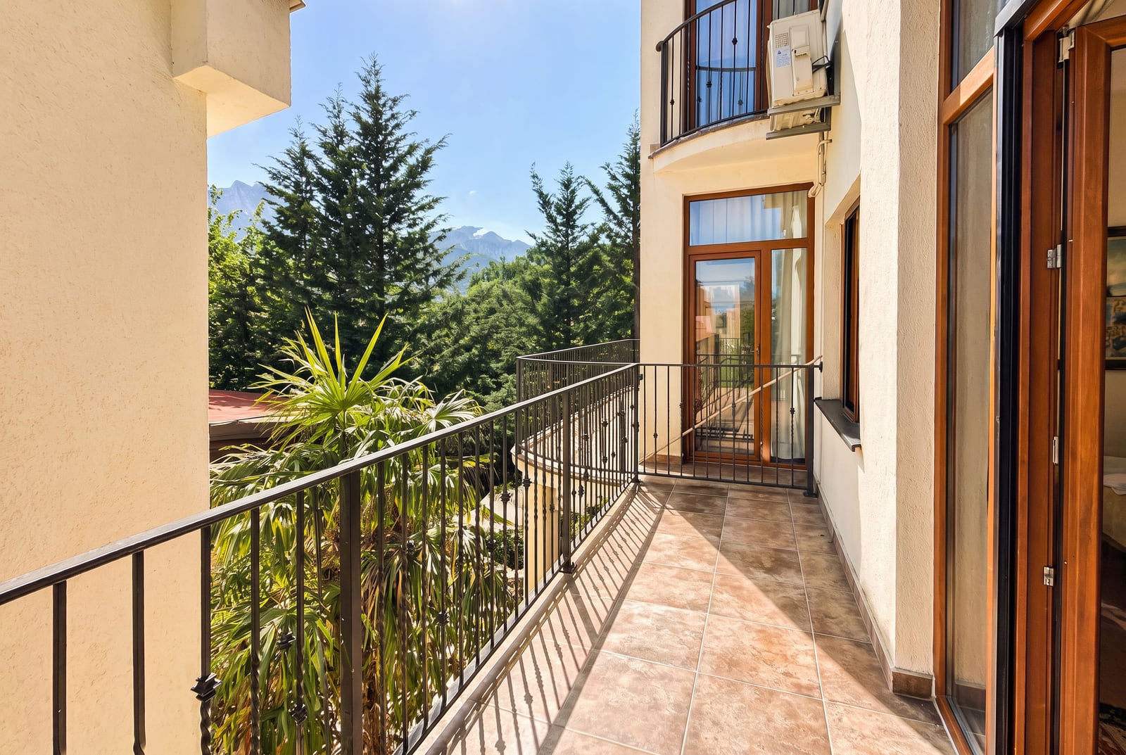 Tiled balcony with black metal railing overlooking trees and mountains, featuring wooden doors and exterior wall.