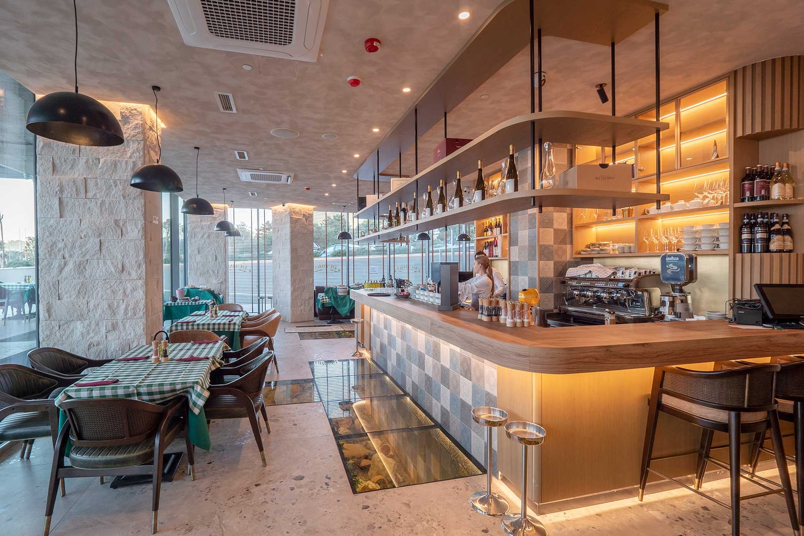 Restaurant or bar interior featuring a wooden counter, bottle shelves, checkered tablecloths, and a glass floor aquarium.