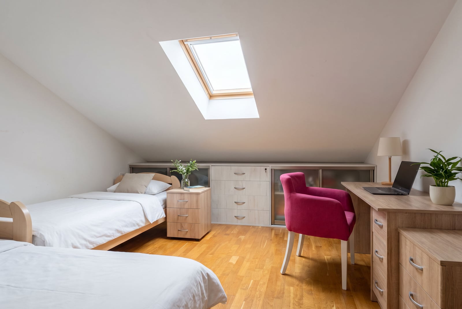 Bright attic bedroom with two beds, a wooden desk, a pink chair, and a skylight in the sloped ceiling.