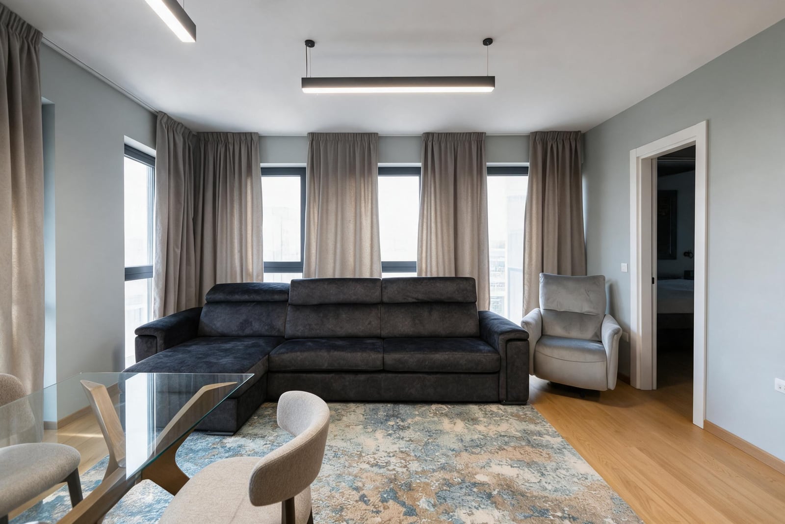 Modern living room featuring a dark gray sectional sofa, glass dining table, patterned rug, and large windows with curtains.