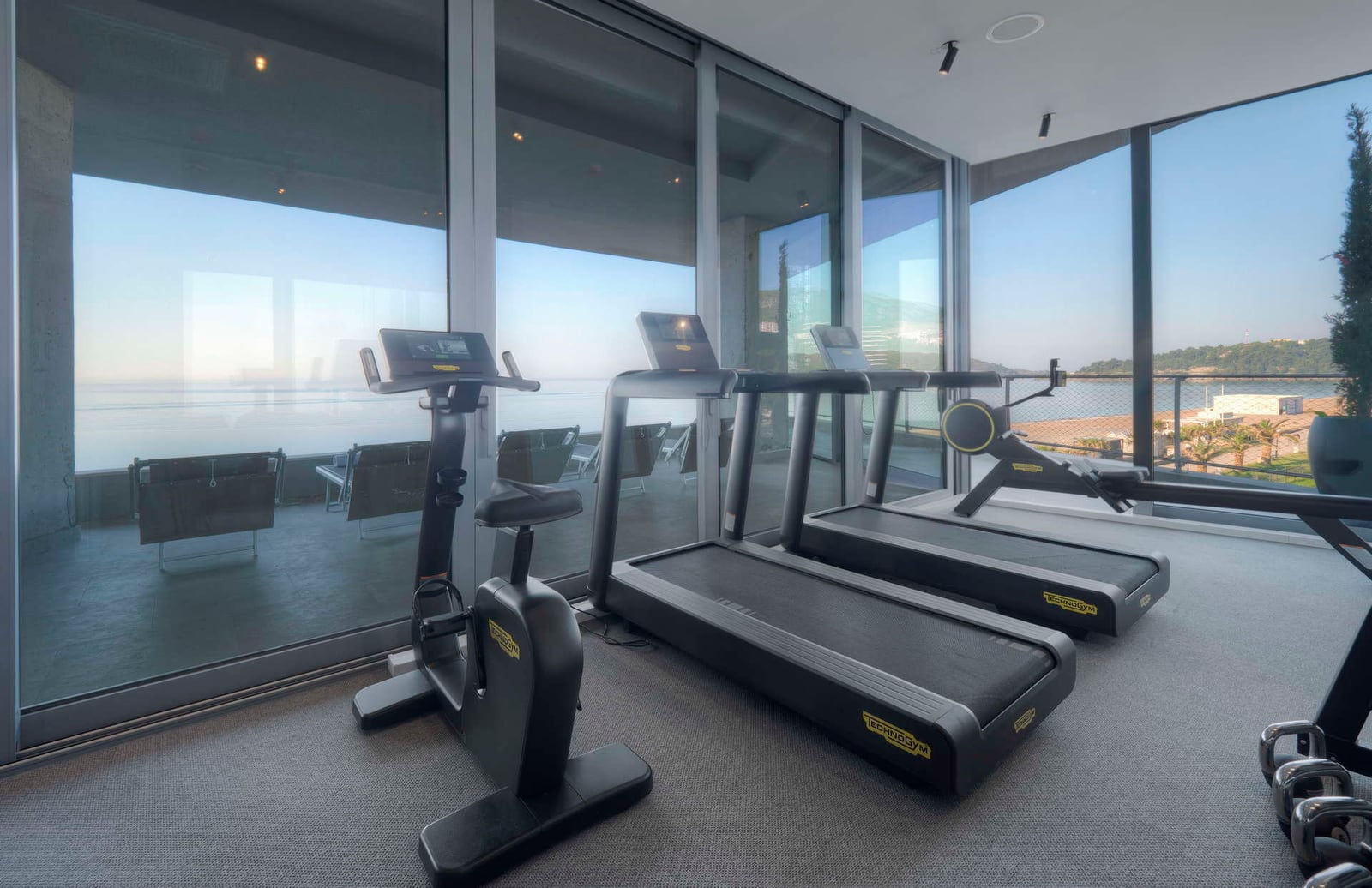 Modern fitness room featuring Technogym treadmills and a bike by floor-to-ceiling windows overlooking the sea.