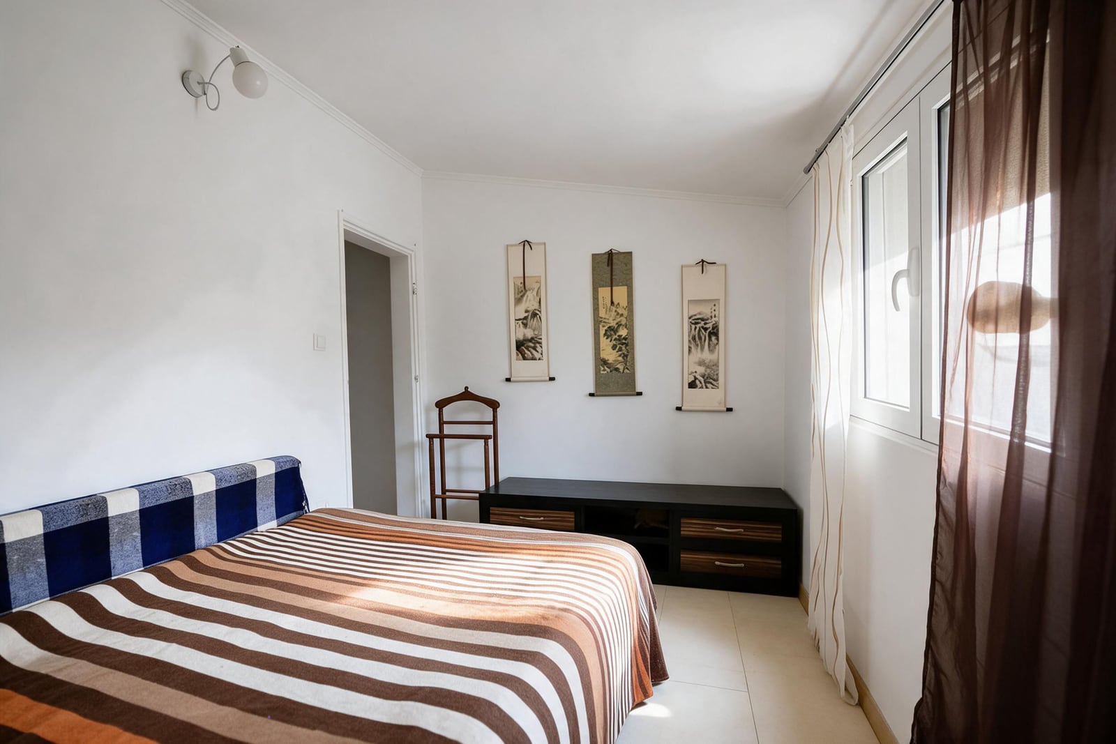 Bedroom with a striped bedspread, black storage unit, wooden rack, and three hanging wall scrolls.