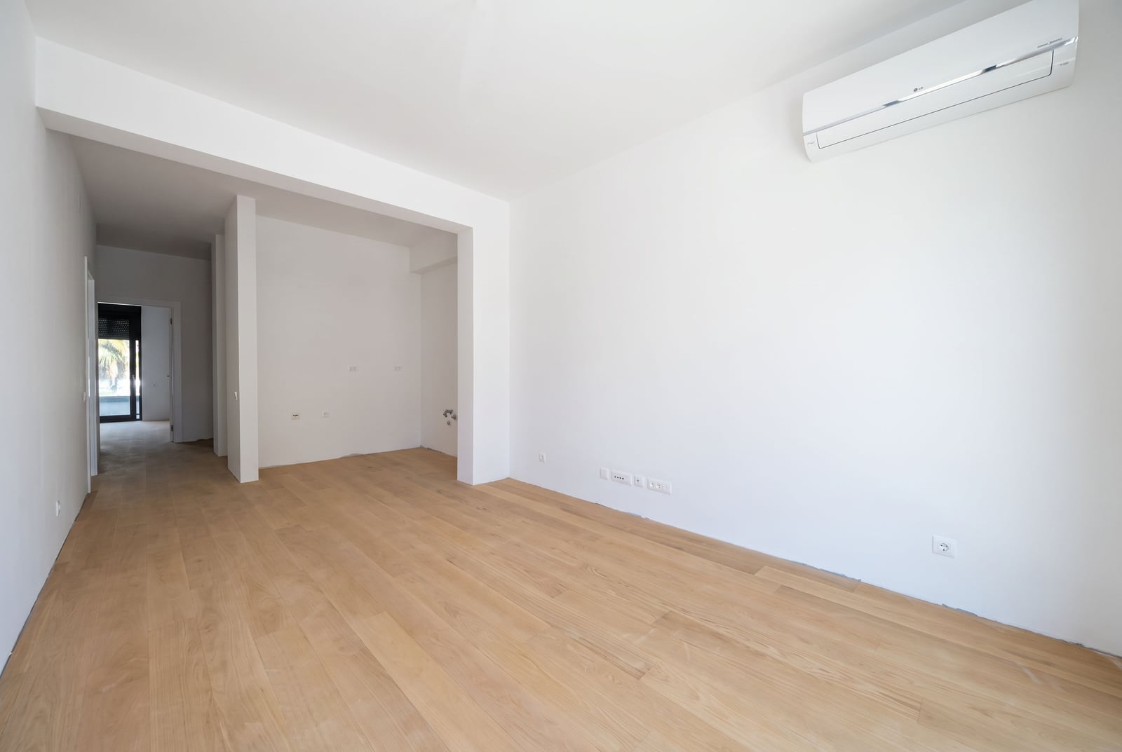 Empty room with light wood flooring and white walls, wall-mounted AC unit, doorway leading to a hallway.