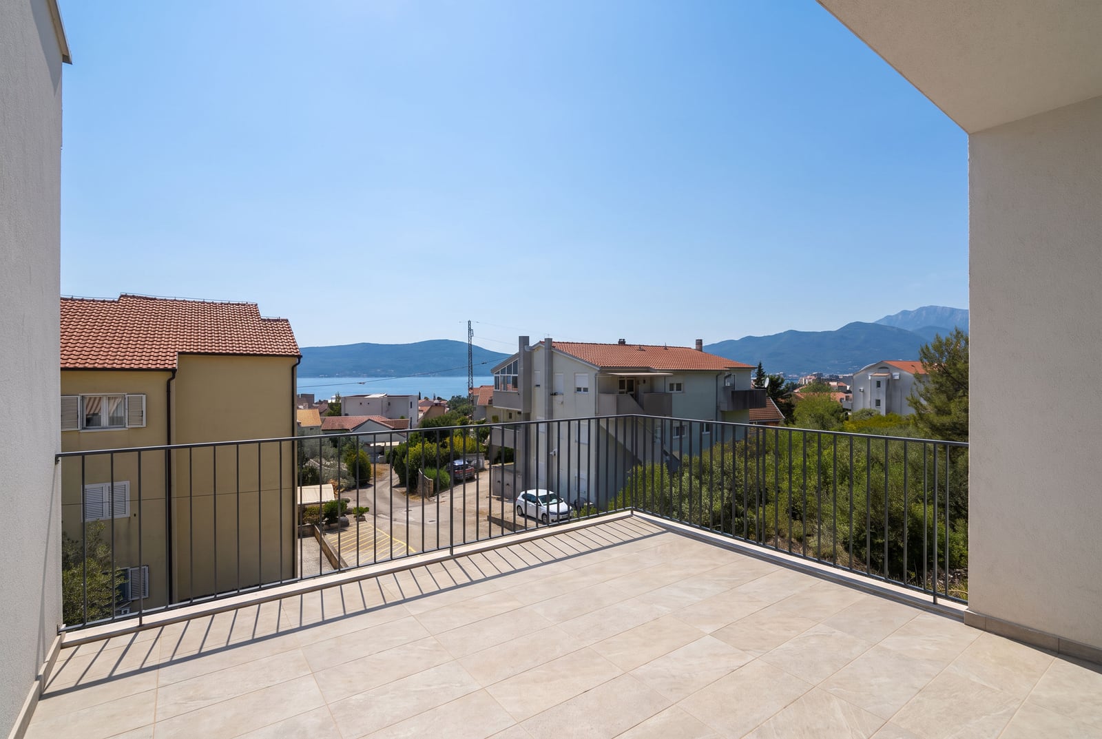 Tiled balcony with metal railing overlooking houses, sea, and mountains under blue sky.