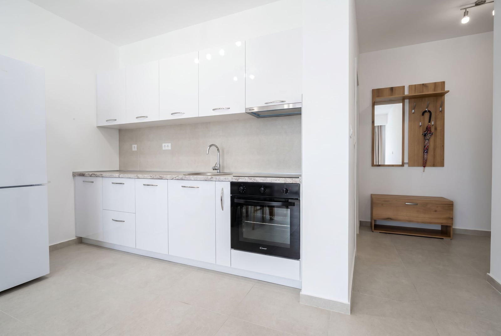 Modern kitchen featuring glossy white cabinets, a black oven, sink, and refrigerator on light tiled flooring.
